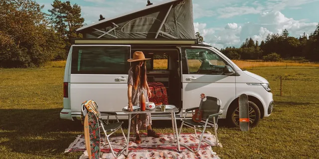 Eine Frau deckt vor einem offenen Camper, der auf einer Wiese steht, den Campingtisch.
