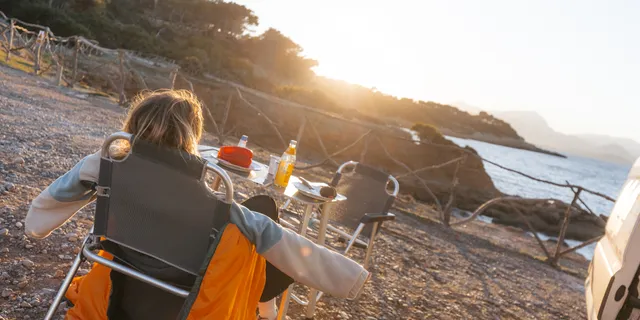 Camper newcomers can relax with camping tips: Like the young woman who looks at the sea in the camping chair in front of a table in the sun and stretches the arms to the side.