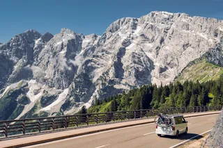 A white campervan with bicycles drives a road, in the background you see a mountain chain, forests and blue sky