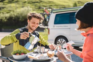 A man with a green sports jacket gives his cup at the camping trip by bicycle at the camping table and laughs at it, facing a woman who melts jam on her bread, in the background a camper with bicycles