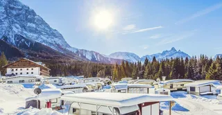 The sun shines on the snowy camping resort Zugspitze. You see some campers and a cabin.