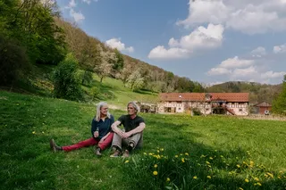 Two people sit on a meadow with wine glasses.