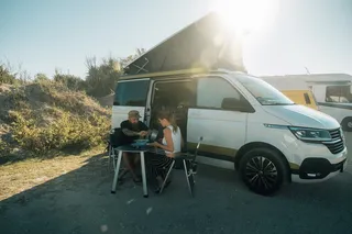 A man and a woman sit on a camping set in front of a camper.