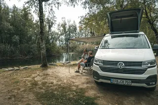 Two people sit in front of a camper parked on a river bank.