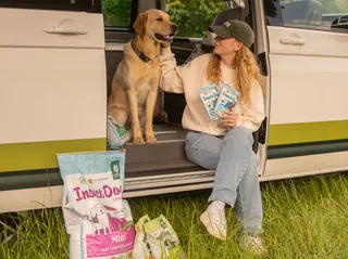 A woman and a dog sit inside a camper on the sliding door. In addition to the camper and in the hand of the woman, dog food from Green Petfood can be seen.